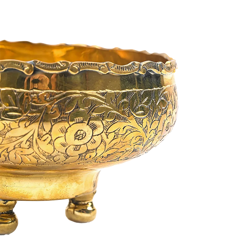 Close-up of a polished brass bowl with intricate floral engravings and three small feet, shown against a plain white background.