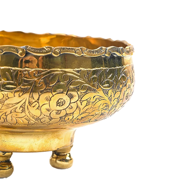 Close-up of a polished brass bowl with intricate floral engravings and three small feet, shown against a plain white background.